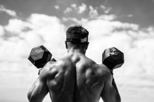 Picture of shirtless male athlete facing away from the camera with dumbbells on his shoulders.