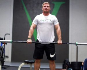 Male athlete standing with an unweighted barbell at his hips.