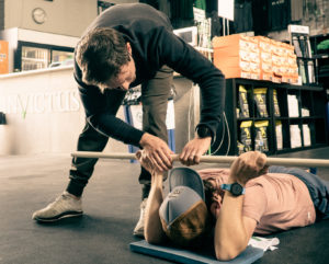 Coach helping an athlete stretch his shoulders with a PVC while face down on floor.